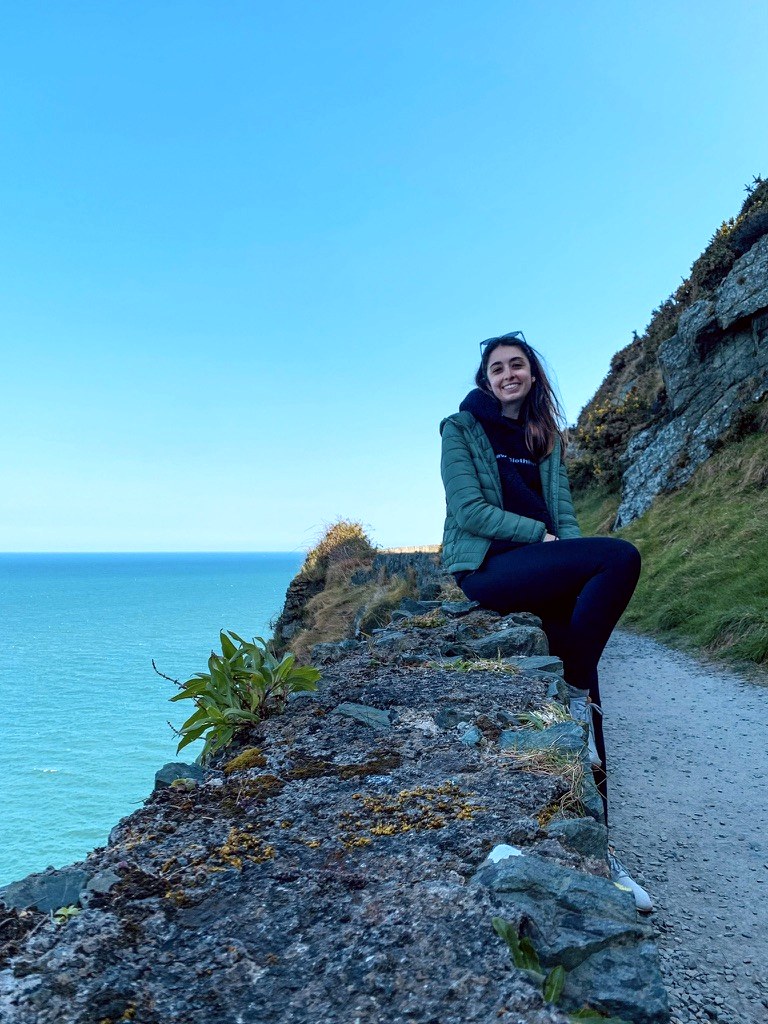 mari staüb sentada num muro de pedra, com o mar atrás
